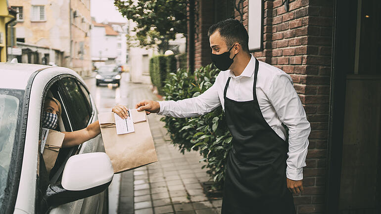 Waiter wearing protective face mask is giving disposable package with food to pretty female driver outside a restaurant