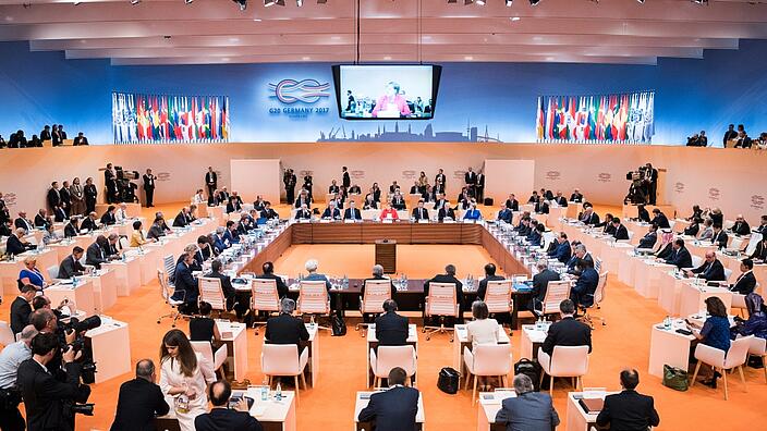 Overview of the G20 Summit 2017 in Hamburg. Angela Merkel is speaking in front of a great hall full of people sitting at their desks.