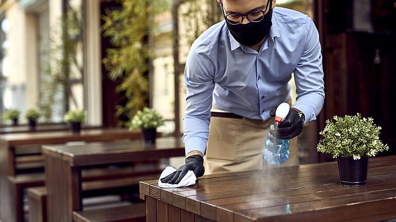 Vorbereitung für die Kunden nach wiedereröffnung / Waiter wearing protective face mask while disinfecting tables at outdoor cafe